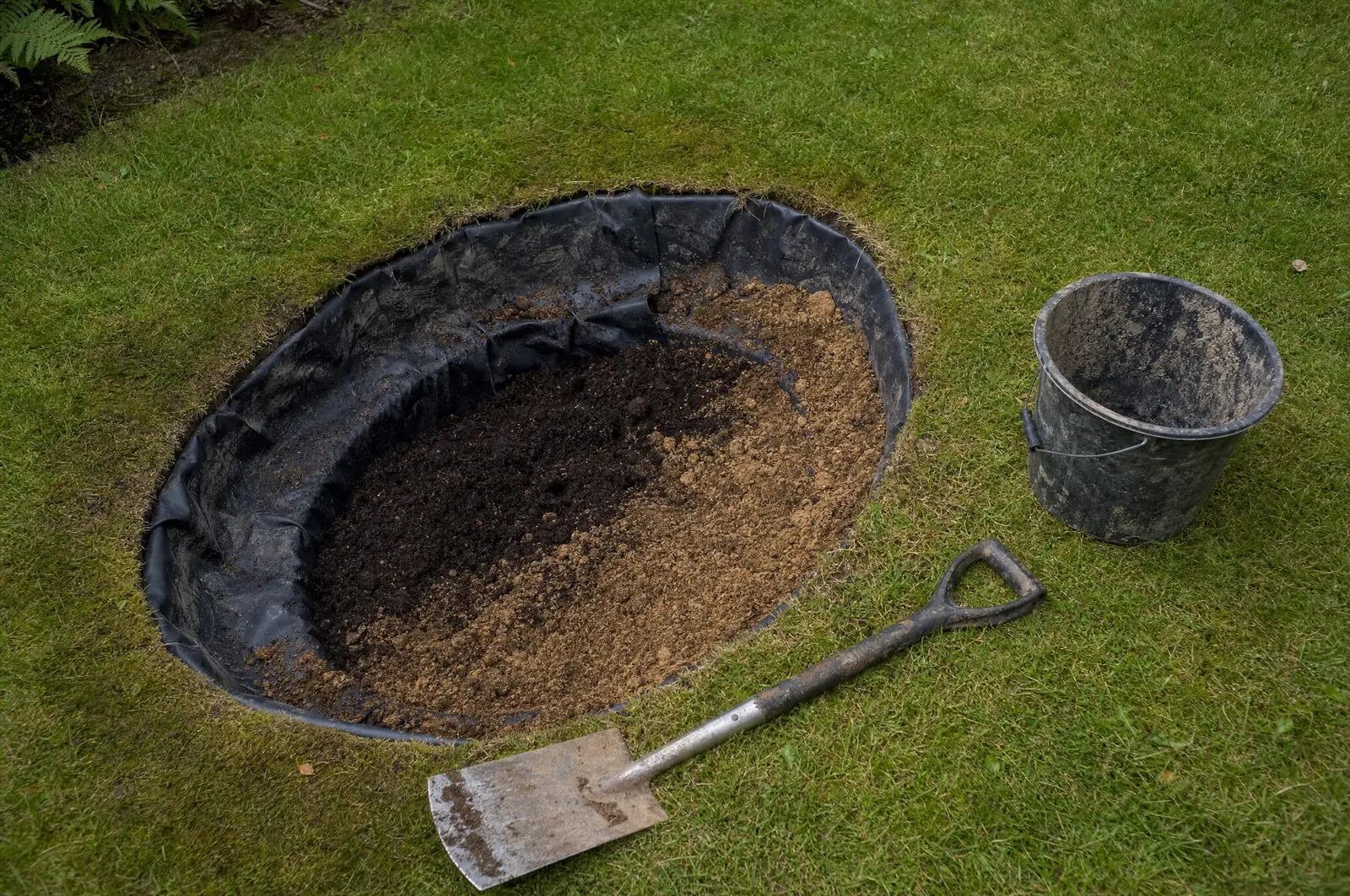 Ovale Grube im Rasen, mit schwarzer Teichfolie ausgekleidet und teilweise mit Torf-Sand-Substrat gefüllt, daneben Spaten und Eimer